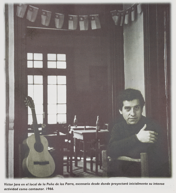 On the righthand side of this black-and-white photo a white man with dark hair wearing a dark sweater looks intently at the camera. He is the singer Víctor Jara, sitting on a chair, leaning his elbows on the back of the chair, arms folded, one hand showing. A guitar is near him, leaning against a wall or doorway. In the background, there are several tables with bottles on them, chairs at the tables, a window, and small Chilean flags strung on a banner that goes across the ceiling.