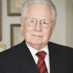 This image shows a white man with white hair, wire rimmed glasses, in a dark suit, red-and-black striped tie, and white shirt looking at the camera. It is cropped to just show his head and shoulders and a little bit of his chest.