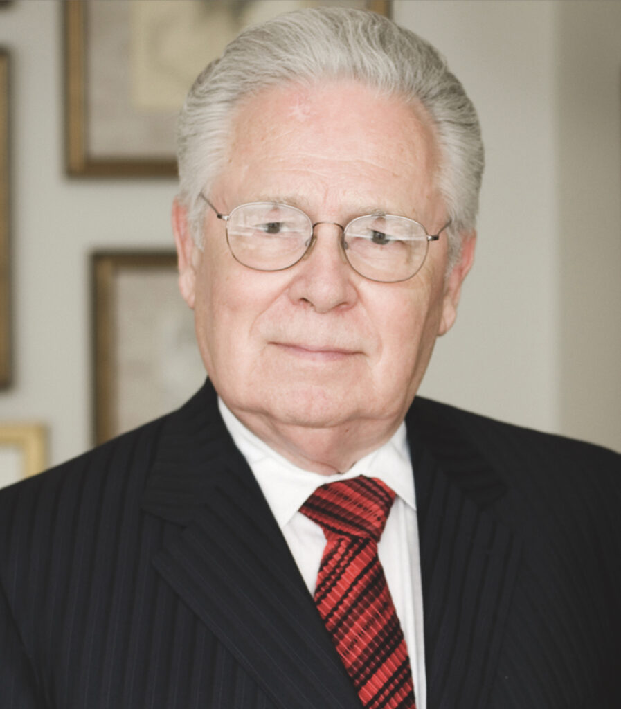 This image shows a white man with white hair, wire rimmed glasses, in a dark suit, red-and-black striped tie, and white shirt looking at the camera. It is cropped to just show his head and shoulders and a little bit of his chest.