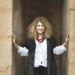 Photo shows author Gioconda Belli, a woman with blondish curly hair just past her shoulders, standing in a stone enclave. Her hands touch the walls on either side of her. She looks directly at the camera, smiling. She is wearing a white shirt, a black short-sleeved jacket and pants, and a large, bright red beaded necklace.