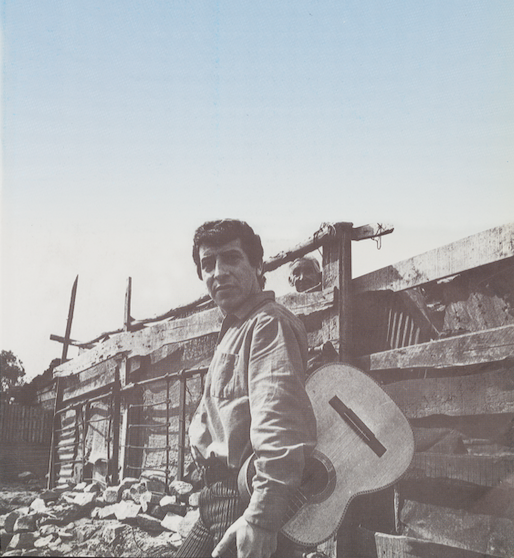 A man stands sideways, outdoors, in front of an old and decrepit-looking fence that might also be the wall of a roughly made house. There are large stones on the ground in the background along with wall. The man, Víctor Jara, looks at the camera. He has a guitar slung over one shoulder. The photo is mostly black and white, but the sky slowly fades to blue above him. In the background, an older man with white hair peers over the fence. We only see his head.