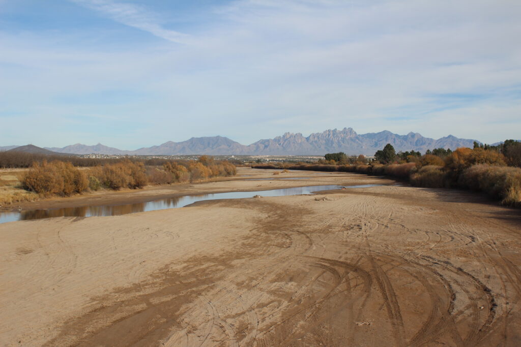 In the foreground, flat, sandy ground with tire tracks. In the middle ground, a small, shallow stream of water crosses the area. On either side there are brownish shrubs. In the middle distance dark green trees are visible. Far in the distance, a town. On the horizon, a rocky mountain range and above it a blue and white sky.