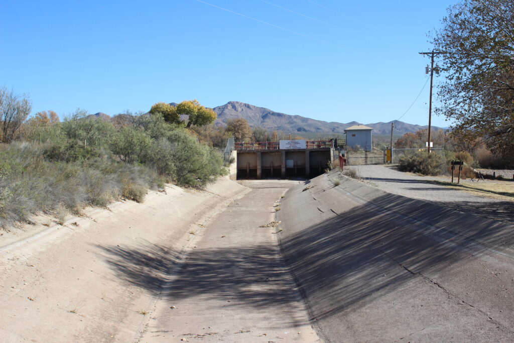 A wide concrete ditch with sloping sides starts at the bottom of the image going toward the middle. A tree from the bank on the right casts a partial shadow over the ditch. On the left-hand bank, scrubby green plants of various kinds. In the distance, a mountain is visible, and above it a blue sky.
