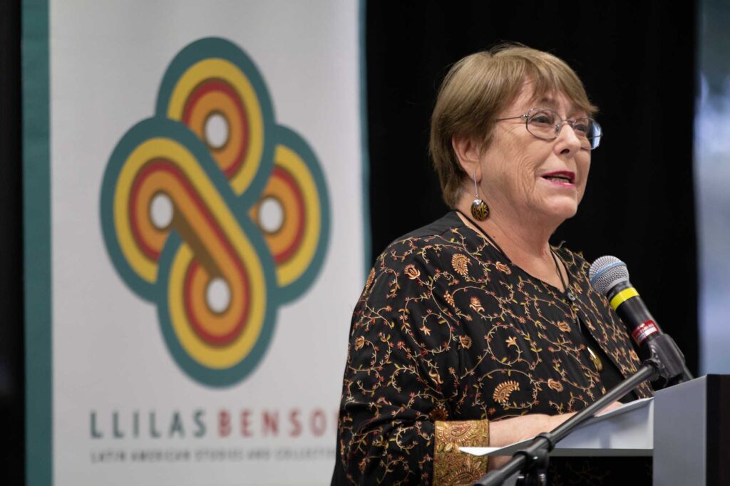 A woman stands at a lectern. She is about 70, with short, auburn hair. She wears wire-rimmed glasses, black-and-gold dangly earrings, and a black jacket with ornate swirling designs in red and gold.