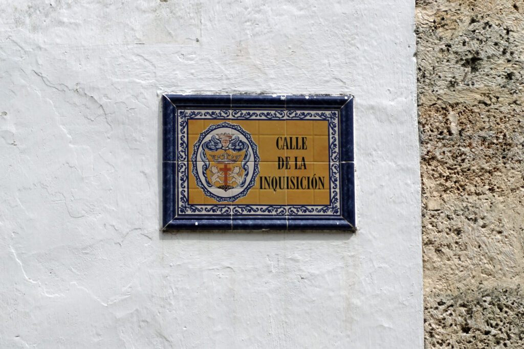 A sign reading Calle de la Inquisición is afixed to a white stucco portion of a wall. The sign is made of tile, with a seal that shows a crucifix, two lions, and a crown. The tiles are goldish yellow, the inner border is ornate blue and white, and the outer border is dark blue.