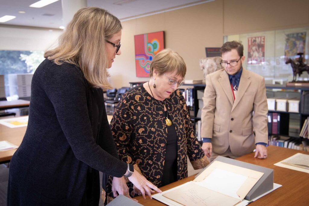 Three people stand at a table and look at an open folder. In the background there is a painting on the wall, some posters, a statue on a shelf, books and archiving boxes on shelves.