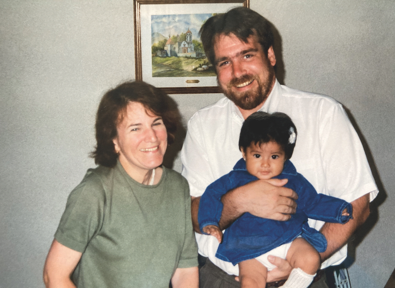 A white woman with brown hair and a sage-green top stands next to a taller white man in a white shirt, brown hair, mustache, and beard. The man is holding a brown-skinned baby wearing a blue dress, with dark-brown hair and very dark eyes. The adults are smiling happily.