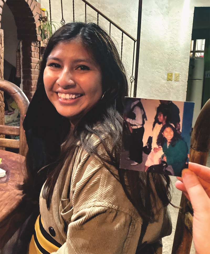 A young woman with long dark hair, dark eyes, and brown skin smiles happily, her head turned to face the camera. To the right of her face, a hand is visible holding a snapshot in which we see several people, including another woman smiling at the camera.