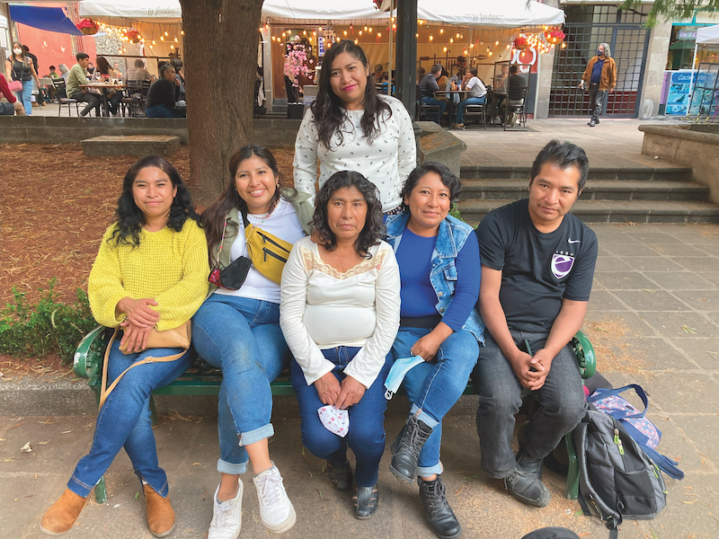 Five adults sit on a green metal bench in an outdoor plaza. One adult woman stands in the center behind them. In the center of the seated adults is a woman who is older than the rest. All but one of the adults are women. They all resemble each other, with dark hair, brown skin, and some common facial features. Some are holding face masks in their hands.