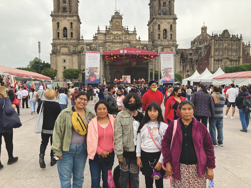 A group of women and girls faces the camera. The girl in the middle is wearing a black face mask. In the background, there is a large, ornate colonial-era church, as well as crowds of people. The white and red tops of tents are visible, indicating that there is a market.
