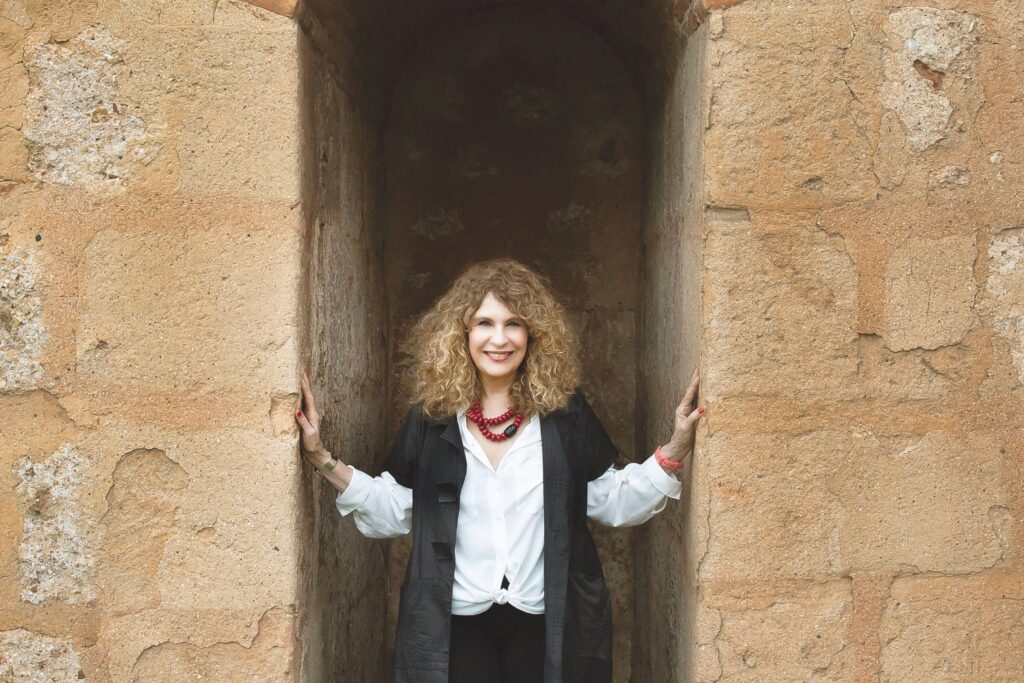 Photo shows author Gioconda Belli, a woman with blondish curly hair just past her shoulders, standing in a stone enclave. Her hands touch the walls on either side of her. She looks directly at the camera, smiling. She is wearing a white shirt, a black short-sleeved jacket and pants, and a large, bright red beaded necklace.