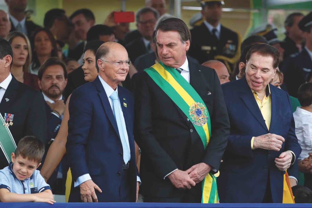 Former Brazilian president Jair Bolsonaro stands in a dark suit with a sash emblazoned with flag colors green and yellow. He leans to the left to speak to a shorter, bald man, also in a suit. On his other side is famous TV personality Silvio Santos, in a dark suit and open-collared yellow shirt. There is a large crowd behind the president.