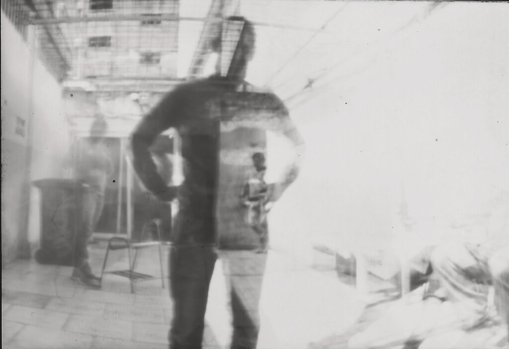 A slightly blurry gray, white, and black image shows the figure of a man in the foreground. He is semi transparent and through him on the right we see another man in the background. On the right, there is mostly bright white. On the left, a gray and shadowy scene shows a man standing near a trash can, a step stool in front of him. The image is a pinhole photograph taken in a prison facility in Argentina by incarcerated people.