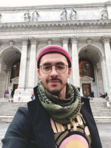 A man in a red cap, round dark-rimmed glasses, an olive-green keffiyeh scarf, and a dark coat looks at the camera. He has a mustache and small trim beard. Behind him is the imposing building of the New York Public Library with its ornate columns.