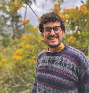 A smiling white man with dark brown curly hair, close shaven mustache and beard, and dark-rimmed glasses looks at the camera. He is wearing a sweater with llamas on it in purples, dark teal, and fuchsia. In the background there are trees and greenery with bright orange flowers.