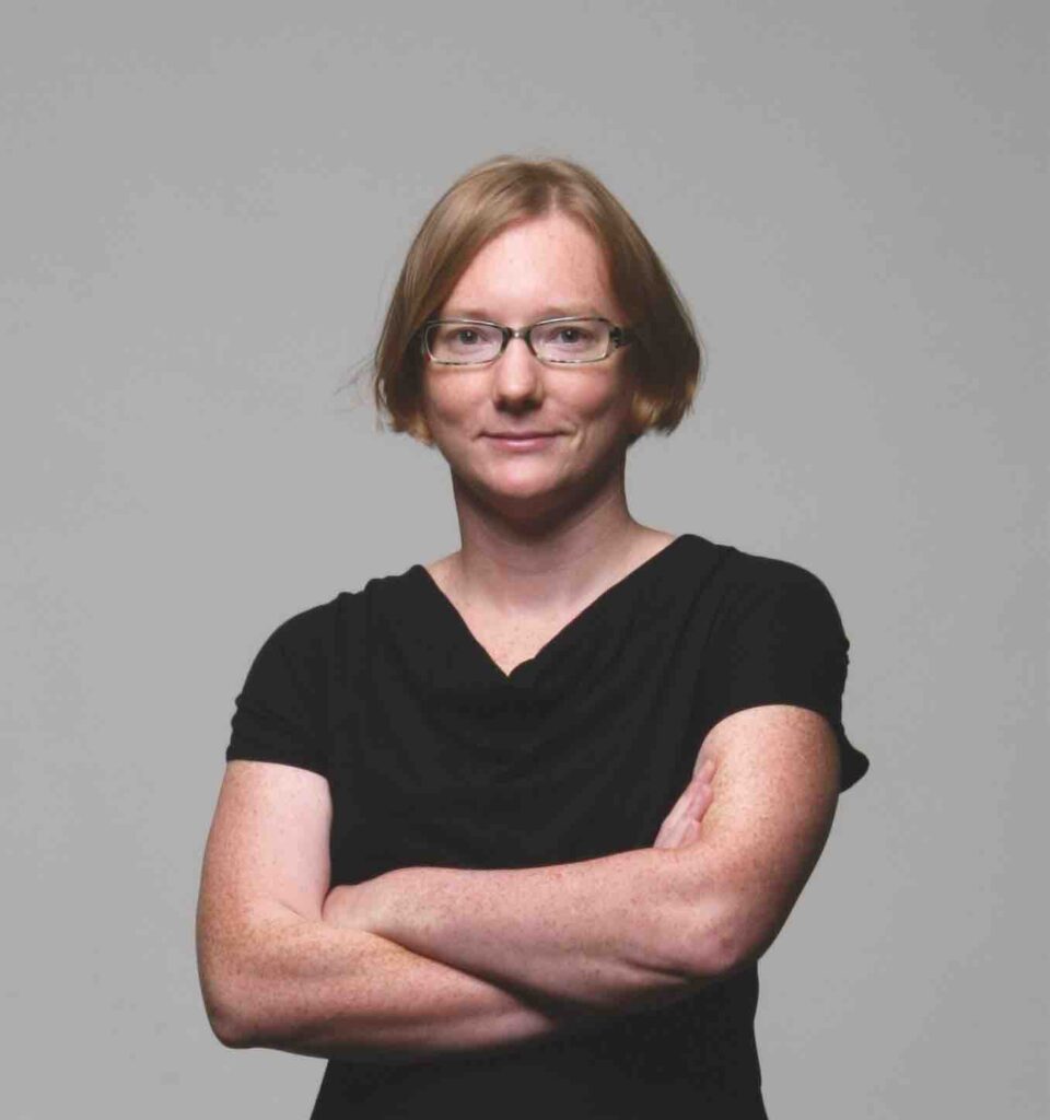 Megan Raby is a white woman with straight red hair that comes to the bottom of her ears. She is wearing glasses, a black short-sleeved shirt that comes to a slight V under her neck, and a small smile.