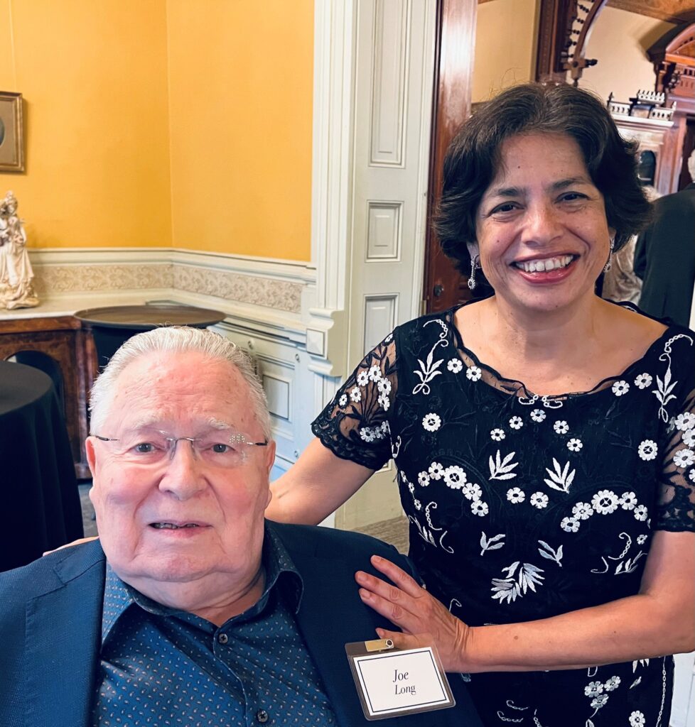 An older white man with white hair, blue shirt and blazer, and wire-rimmed glasses is seated. Standing beside him with her hands on his shoulders, and smiling, is a woman with dark-brown hair, medium brown complexion, wearing a black top with white flowers embroidered on it.
