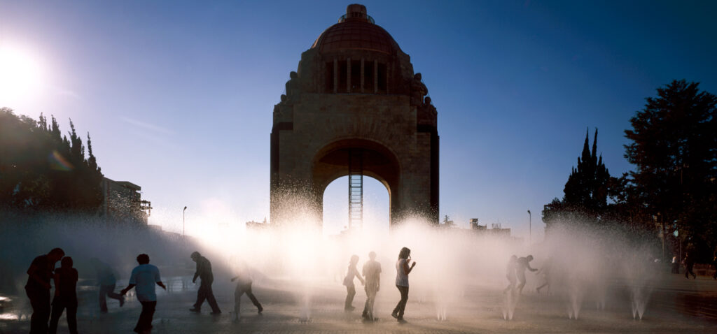 A large arched monument with a domed top is at the center of this photo. Blue sky is behind it. In front, small fountains on the pavement spray mists of water upward. Pedestrians walk among the sprays, backlit by the white misty clouds of water.