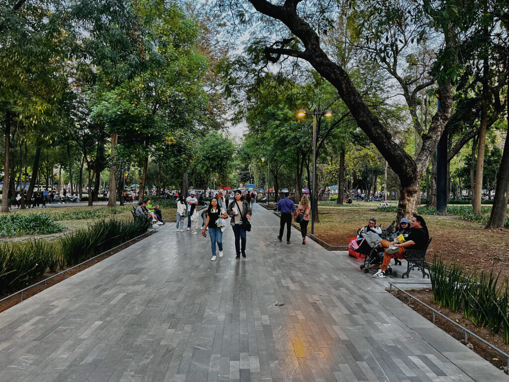 A broad walkway made of gray stone rectangles of different shades is the central feature of this photo. The walkway is in a park and shaded by trees on both sides. People walk along the path, others sit on benches. In the background colorful table umbrellas and many more people are visible.