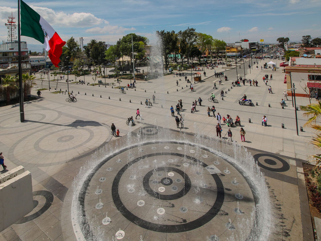 Aerial view of a large, broad plaza and streets open to pedestrians only. In the foreground, a fountain features concentric circles of dark stone and many jets of water coming out of ornate metallic disks. A large Mexican flag of green, white, and red waves from a pole at left. People walk, ride bicycles, and sit on the many pilons that dot the area. A park with trees, low buildings, and eventually a few cars can be seen in the distance.