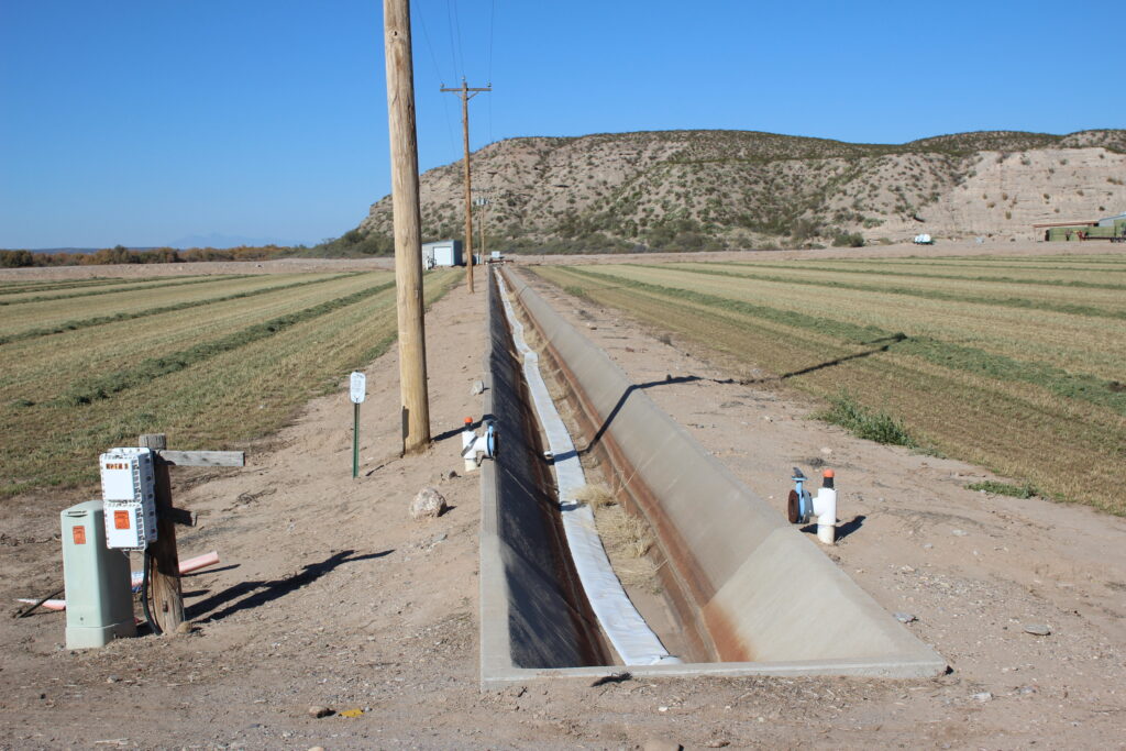 A narrow concrete ditch starts at the bottom of the photo and extends far out. It has a flat white hose lying in it. On either side, the ground is dirt and then short grass in a field of some kind. There are telephone poles along the ditch. In the distance, rising to the right, a brown hill with dark green and grown scrubby plants.