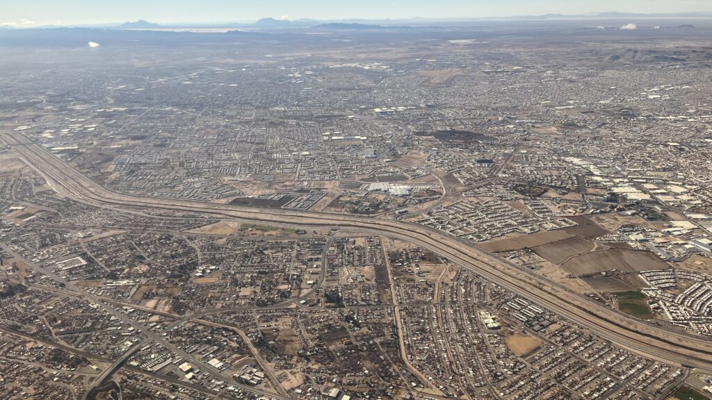 Aerial view of the cities of El Paso and Juárez, traversed by the lightly curved river channel, which appears as a brown series of parallel lines. In the far distance some bluish and grayish mountains are visible under a pale blue sky.