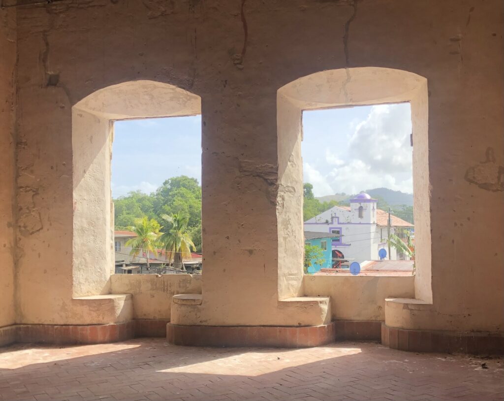Two very large windows cut into thick stucco covered walls look out onto a distant mountain, palm trees and other vegetation, and a few buildings. The windows have no curtains, glass or bars.