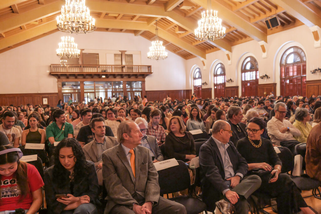 A large crowd of people sits in rows in a room with large windows and four chandeliers hanging from the high ceilings.