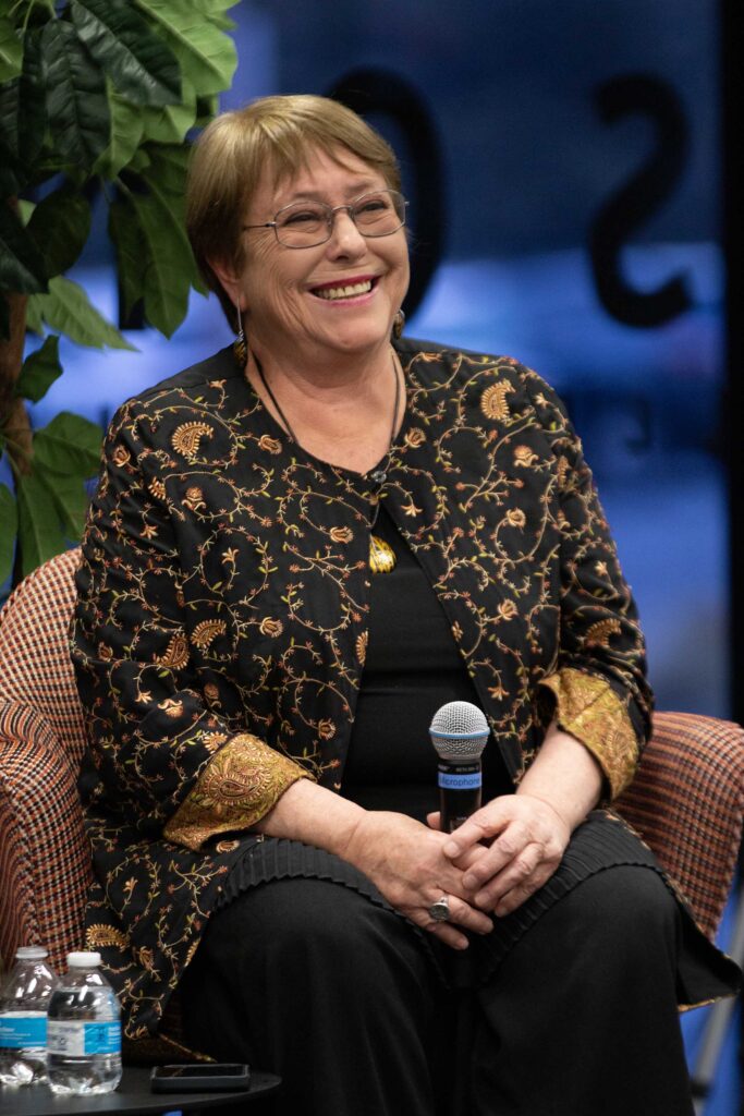 In a close-cropped photo, a woman of about 70 sits in a chair, smiling broadly, holding a microphone near her lap. She has short, auburn hair and wire-rimmed classes, and wears black top and pants with a black and gold jacket decorated with ornate designs.