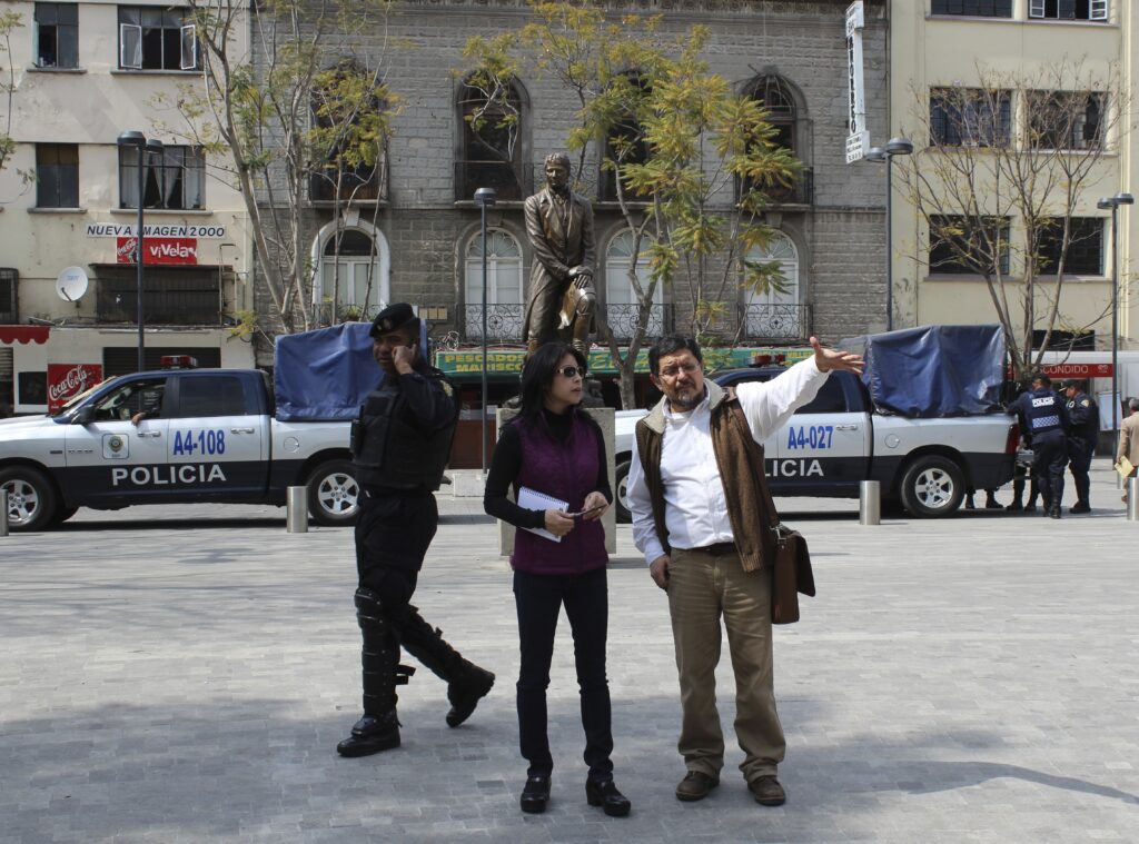 Three people are on a broad sidewalk. In the middle, a woman with dark hair and sunglasses holds a notebook and a pen. She is listening to a man who is gesturing at something with his hand. A police officer walks by the holding a phone to his ear. In the background there two white-and-blue police pickup trucks and more police. Across the street, a row of buildings and a large bronze statue of a man.