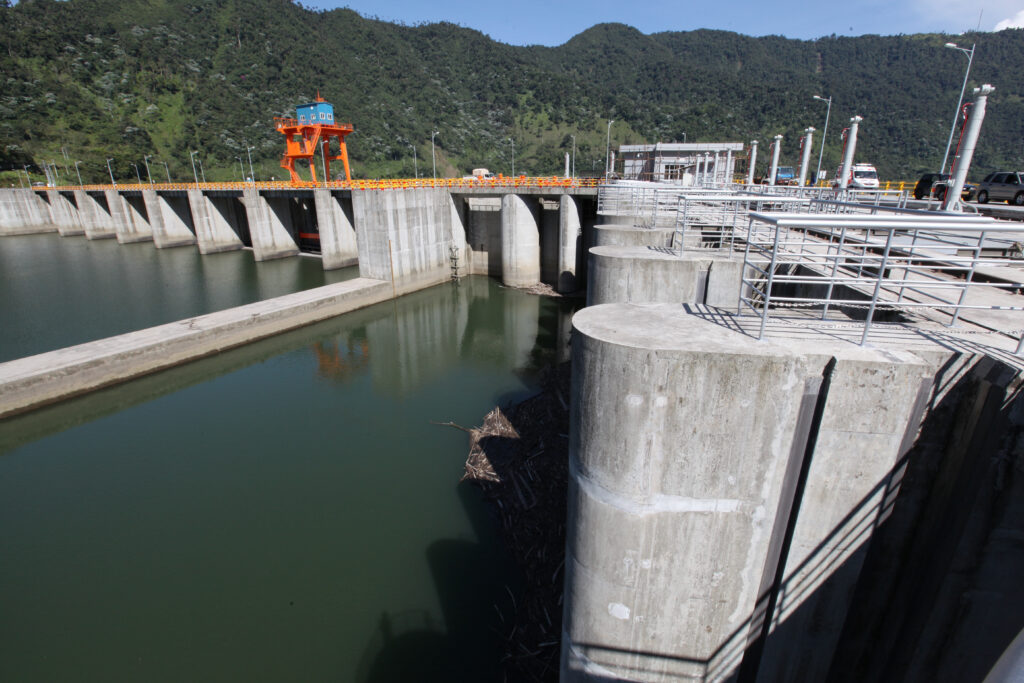 A complex structure rises above a large body of water, with many columns and metal fencing. Along the right a bridge is visible, attached to the structure, with various vehicles on it. There is a bright orange structure rising above everything with a small, bright blue building on it, like a watch tower. In the background, rolling green hills.