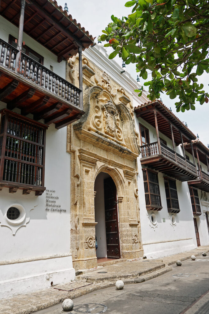 An ornate stone doorway on a two-story white stucco building. The building has wooden bars on the windows and wooden balconies. Letters affixed near the doorway identify the building as the Palace of the Inquisition, Historical Museum of Cartagena.