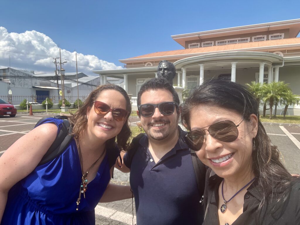 Three people smile and pose for the camera in a parking lot. Two women and a man in the middle. All have dark sunglasses on.