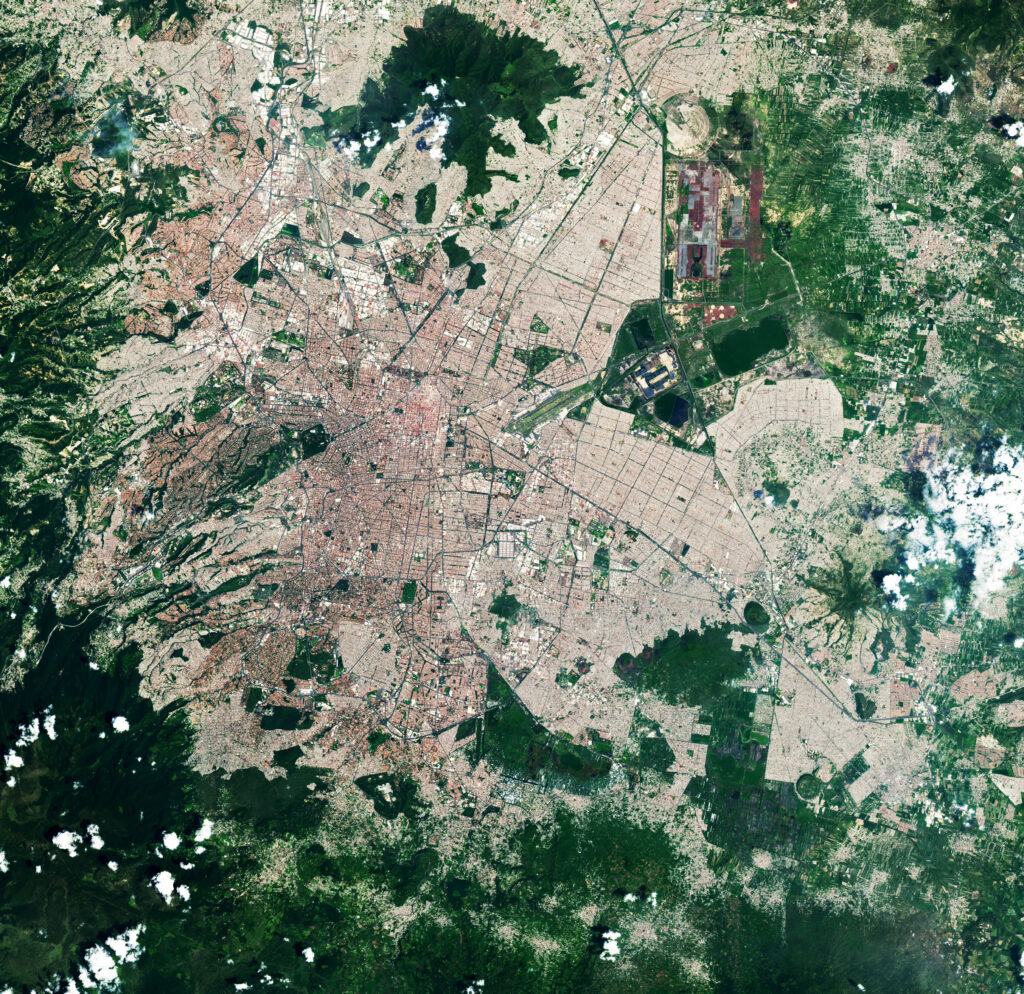 In this satellite image of the Basin of Mexico, there are patches of rich, dark green near the edges of the image and a lot of pinkish and beige areas divided and subdivided into squares and rectangles. These are traversed by straight dark-green lines. Occasionally there are blocks of vibrant pinkish red.