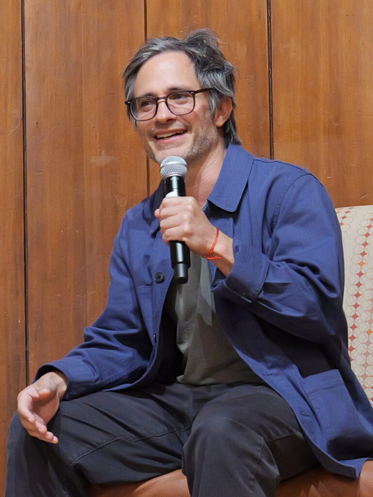 Close-up photo of a seated man with grayish hair, dark-rimmed glasses, a blue jacket, and the shadow of a beard. He holds a microphone and is speaking, a smile on his face.