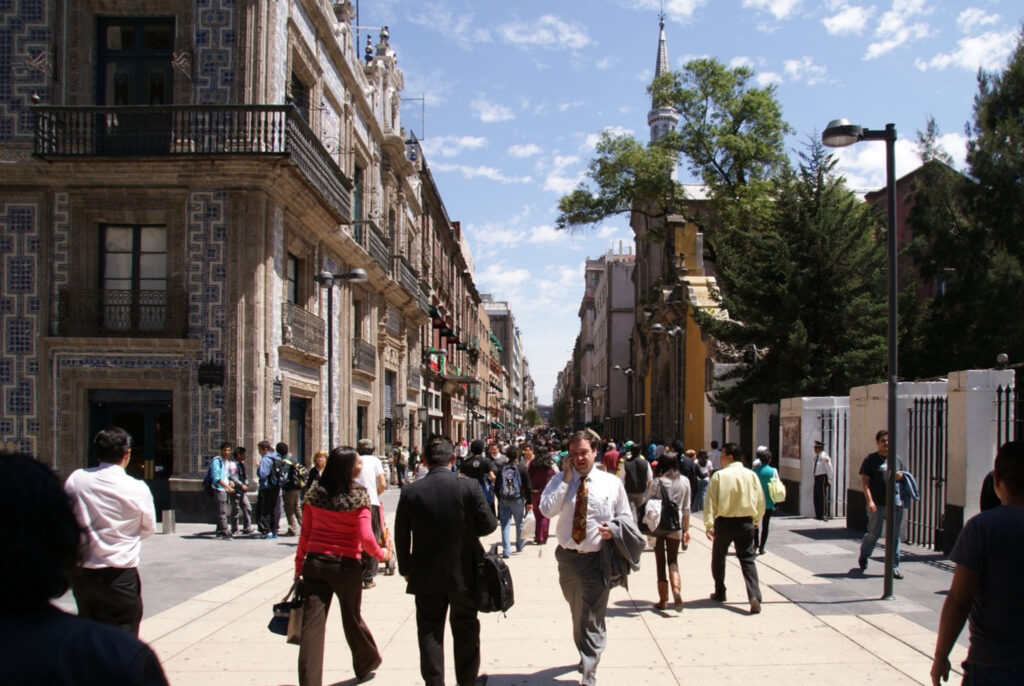 Many people walk on a pedestrian corridor. To the left, and ornate three-story Spanish-era building with blue-and-white tile and wrap-around balconies begins a block of buildings that extend to the vanishing point. On the right, a church steeple rises high above a bright-yellow façade.