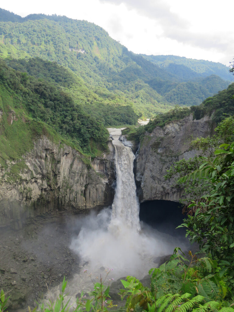 A large waterfall gushes through high rocks amid lush, green mountains.