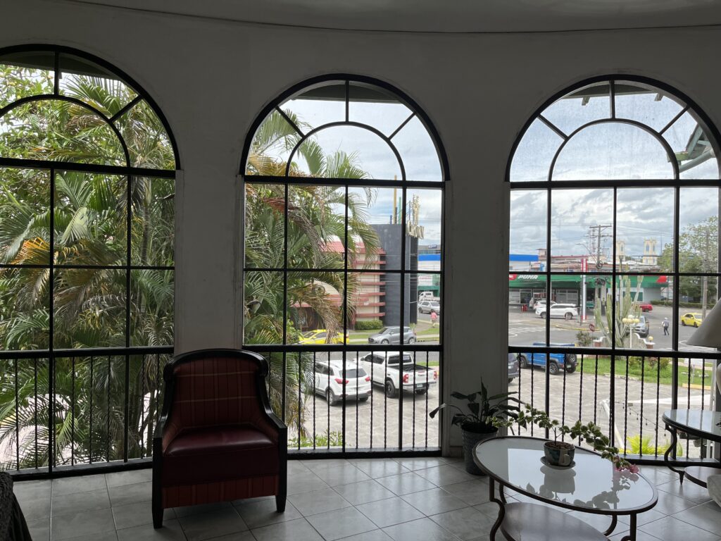 Three floor-to-ceiling windows look out on a large palm tree, a parking lot and some buildings below. The windows are in a sitting area with white tiled floor. They appear to have screens but no glass. They have curved tops and a railing across the bottom.