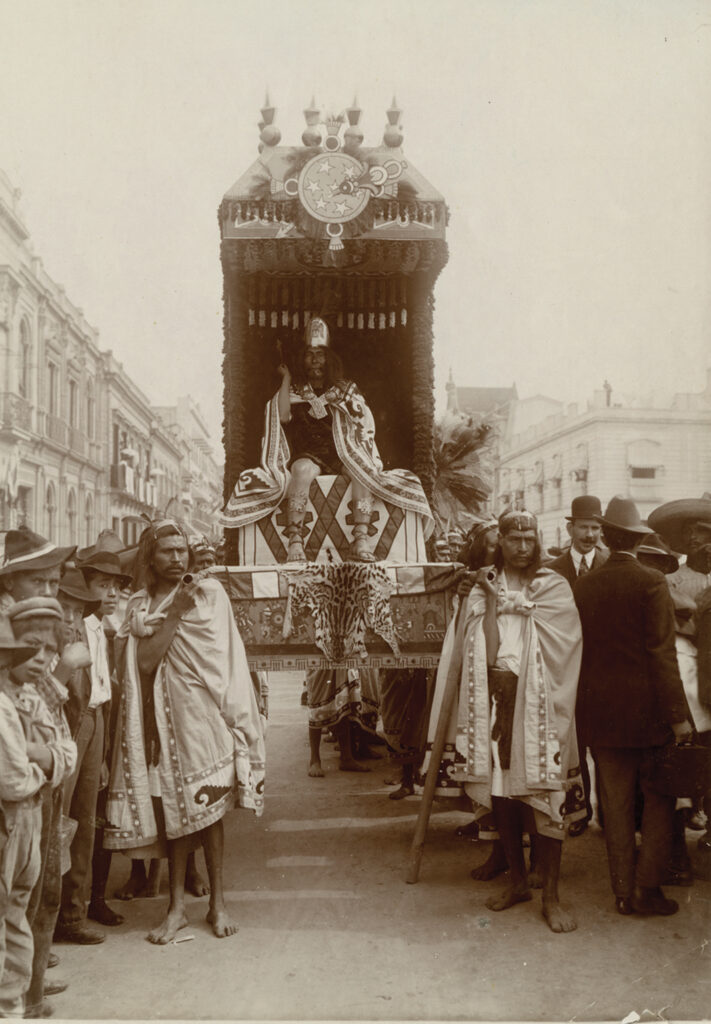 Sepia-toned photograph of a street procession. Barefoot men in capes carry a tall, ornate palanquin with a crowned and robed man sitting on the throne. Parade attendees are visible—children on the left and men in hats and suits on the right.