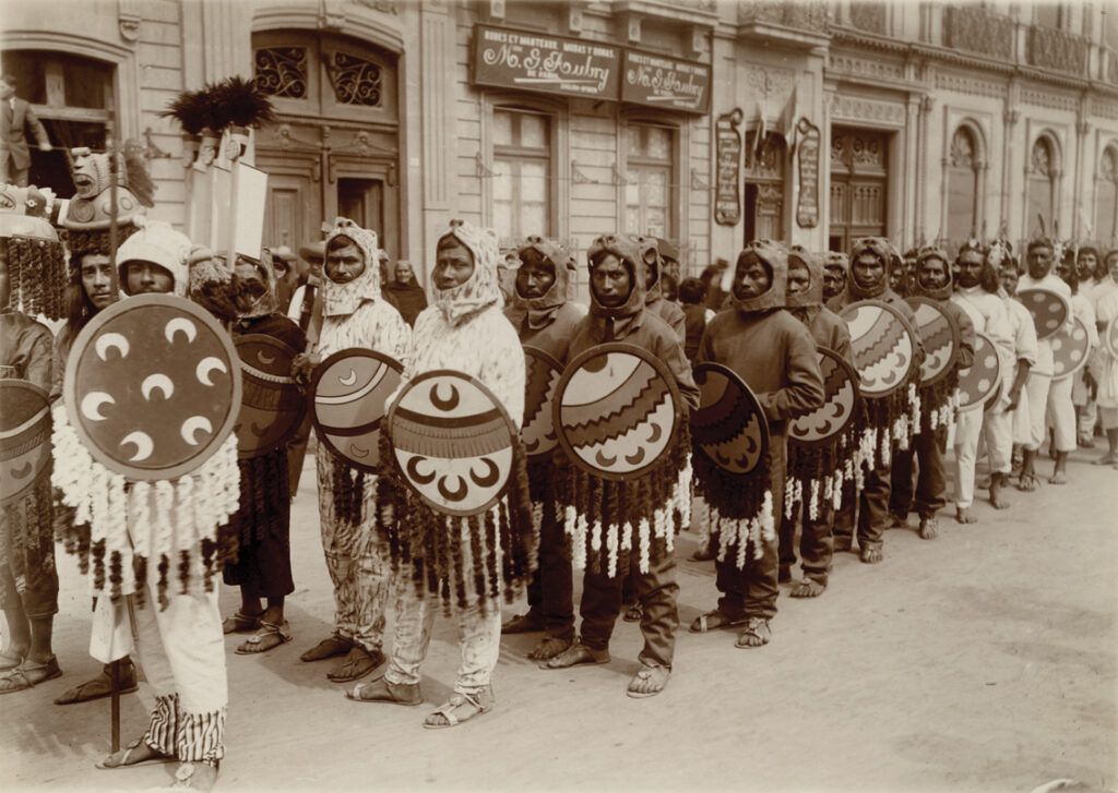 Sepia-toned photograph of rows of men dressed in two different shades of matching outfits that have hoods. The men have dark skin. They are wearing sandals and each one holds a large round decorated shield.