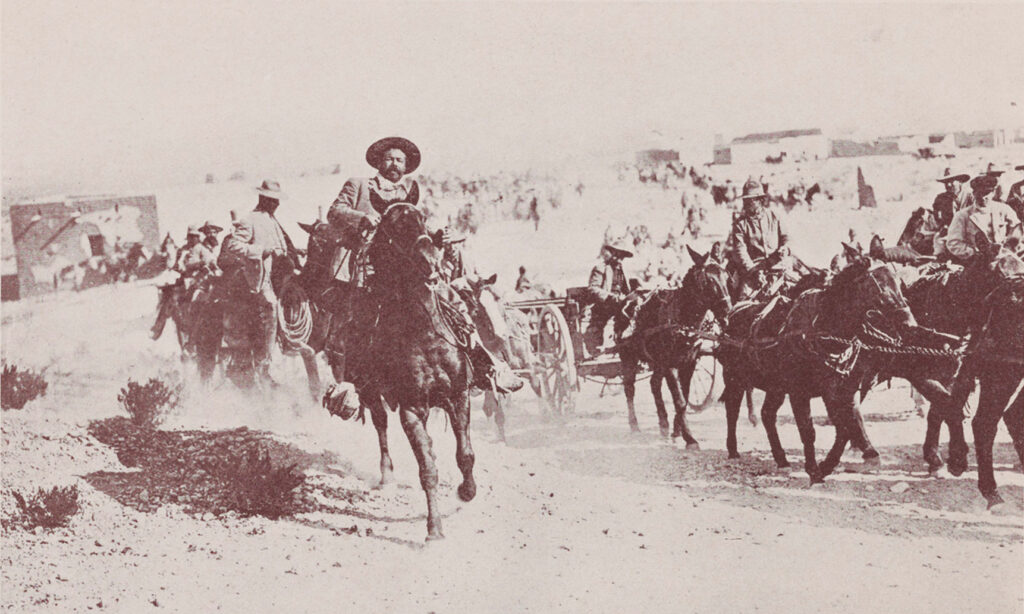 Old black-and-white photograph shows many people on horseback, including some pulling a wagon. At the front near the center is a large man with a round face and a sombrero, the revolutionary leader Pancho Villa.