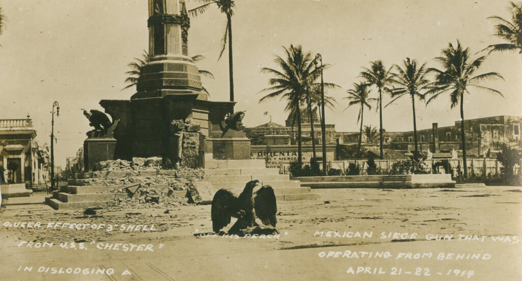 A black-and-white photograph shows a desolate landscape. Front and center is the statue of an eagle, its beak open. Behind that, a partially destroyed monument with other eagles whose stairs and one pedestal are crumbling. Written in white underneath the loan eagel: "Off His Perch." Across the bottom of the photo: "Queer effect of 3" shell from the U.S.S. 'Chester' in dislodging a Mexican siege gun that was operating from behind. April 21-22-1914."