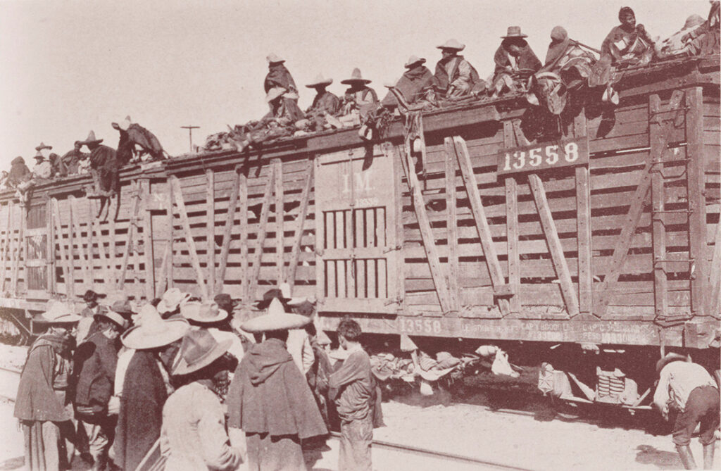 A faded black-and-white photograph shows a wooden train car with many people, mostly women, sitting on the top. Most wear sombreros and are wrapped in blankets. Other people in sombreros and blankets stand in a group on the ground, next to the train car. The car also has what appears to be clothing on the platform underneath is, and at the edge of the photo, a man in boots bends over looking underneath the train.