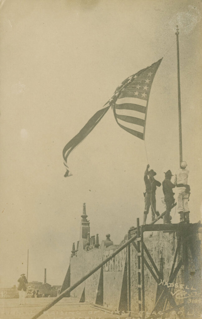 Faded black-and-white photo shows a U.S. flag being raised on a pole by three men in uniform. One is in a white sailor's uniform and the other two wear darker colors. They and the flagpole are on top of a stone fortress wall. There is a partially illegible white transcription at the bottom of the photo. It reads in part "raising of the first flag of U.S."