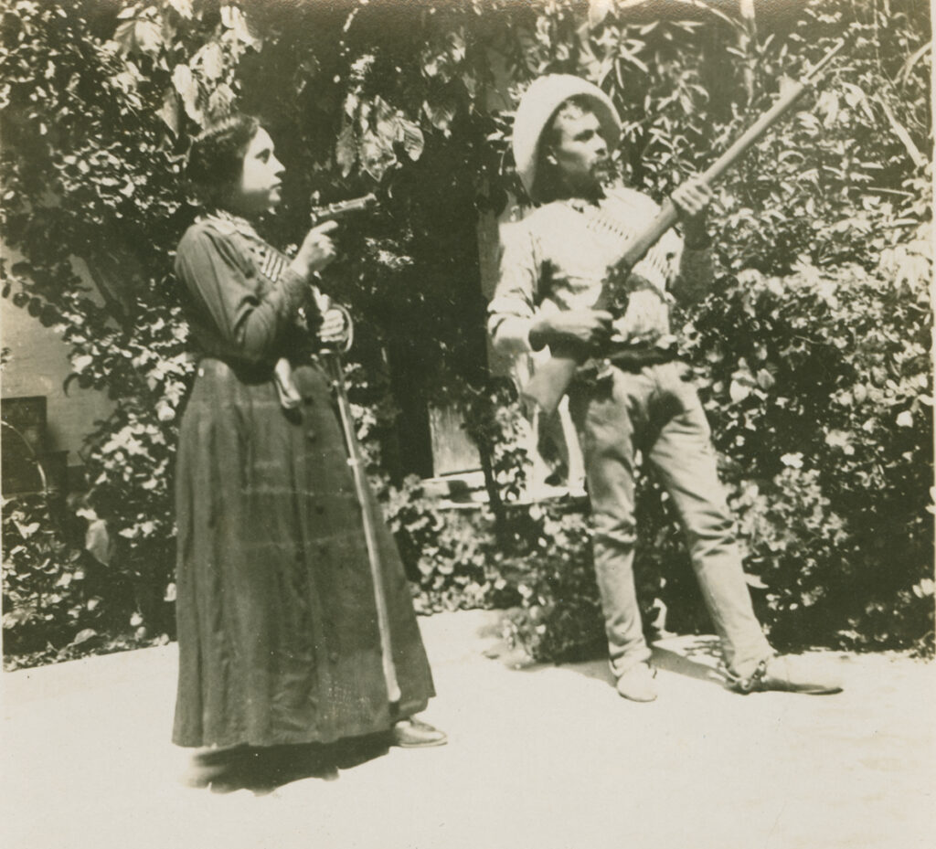 In an old black-and-white photo, a man and a woman face toward the right, both holding guns. The man is wearing a sombrero, spurs, and a bandolier holding bullets across his chest. He holds a long rifle in both hands, which crosses his chest and points upward. The woman is wearing a long dark dress, also with a bandolier across her chest; she holds a small pistol.