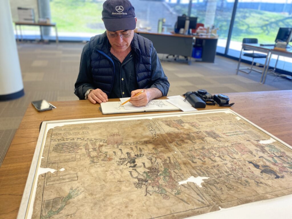 A man in a dark blue cap and dark blue clothing sits at a wooden table and sketches with a pencil. Laid out in front of him on the table is a large and somewhat damaged painting on very old paper.