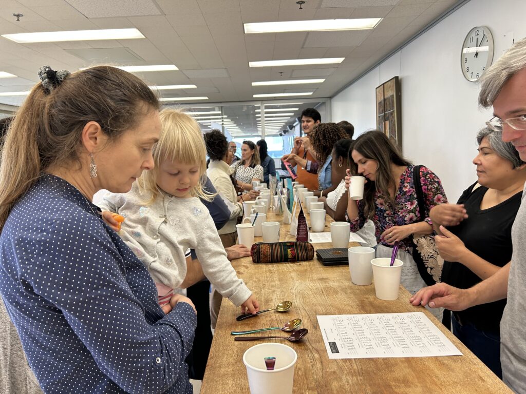 People stand on both sides of a long table with many white cups on it, at a coffee tasting.