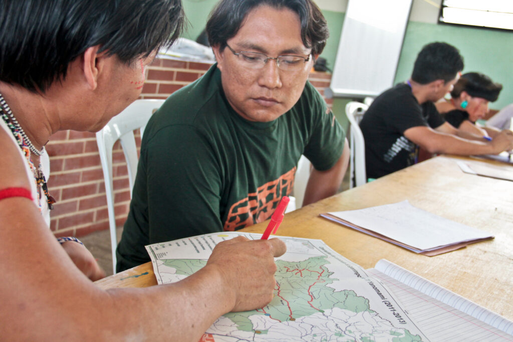 Two Indigenous men sit at a table in the foreground and review a map of Yanomami territory (2011-2013). One wears face paint and beads and holds a red pen. The other is wearing wire-rimmed glasses and a t-shirt. Farther down the table two other men sit. One has a black band on his forehead, red facial markings, and a large turquoise earring. The other wears a t-shirt.