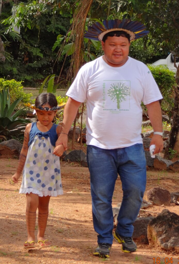 A man with dark hair and a round face walks outdoors on a dirt path holding a young girl by the wrist. He wears a round headdress of blue feathers, a white t-shirt that says Flor do Cerrado, Brasília, Brasil in green, with a green illustration, and blue jeans. The girl has a similar headdress and wears a dress with blue, white, and yellow polka dots, and yellow flip-flops. She has a series of horizontal lines on both legs. Forest plants and grass are visible in the background.
