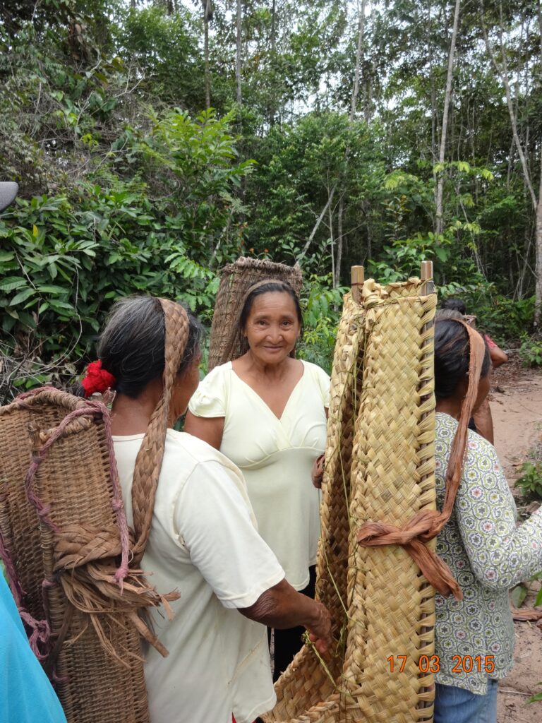 Three women are in a forest. Each carries a large, long woven basket along her back. One is facing the camera. She has a wrinkles, dark hair, and is wearing a cream-colored dress.